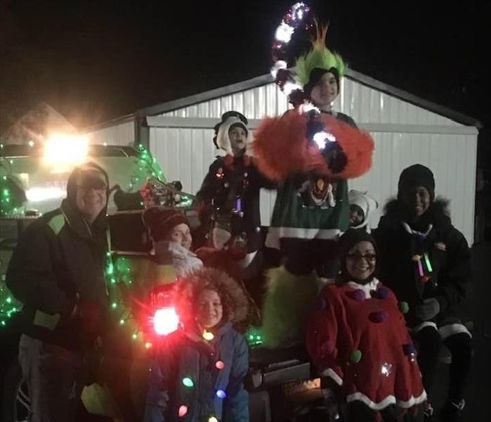 Several SERVPRO employees and children dressed in Christmas sweaters on the bed of a SERVPRO truck decorated with lights